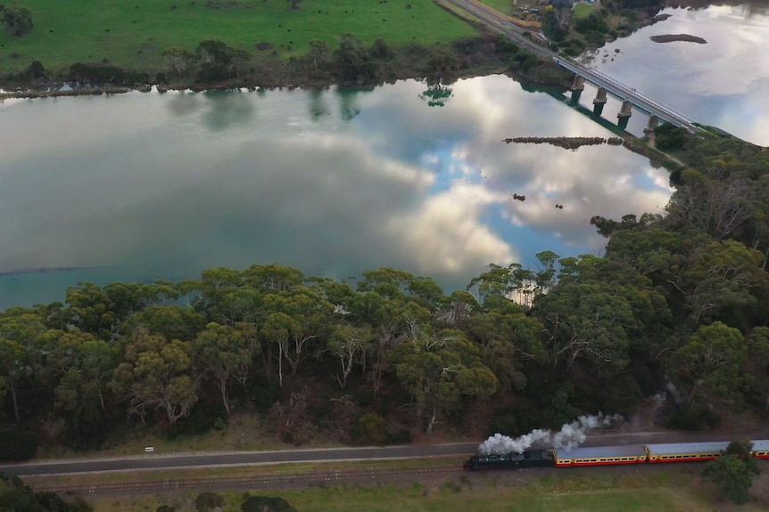 Aerial view of a steam locomotive pulling three carriages close to a serenely still and broad river.