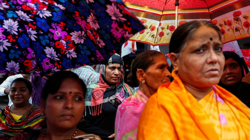 Women in bright saris with serious looks on their faces shelter under floral umbrellas