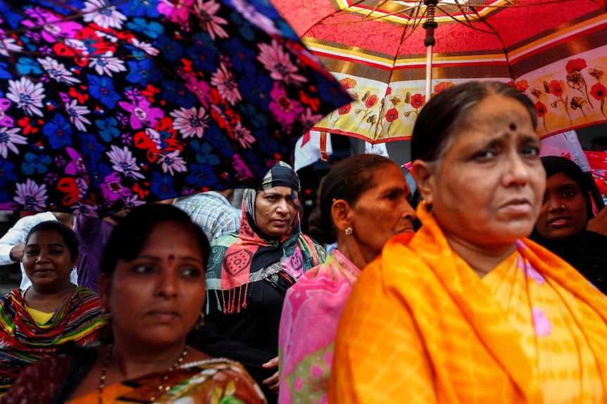 Women in bright saris with serious looks on their faces shelter under floral umbrellas