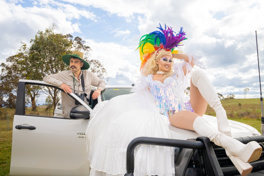 A man in a Bunnings straw hat and a drag queen lean on a white ute.