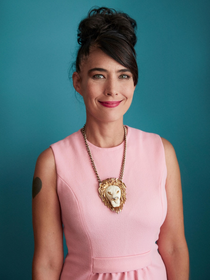 Profile picture of Kathleen Hanna - she has her hair in a bun, is wearing a pink dress and a lion's head necklace