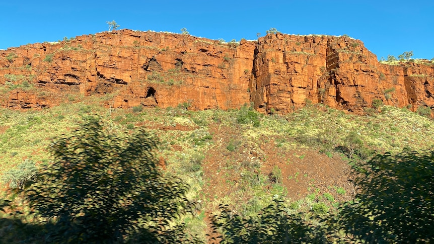 Red cliffs rise above a scrubby hill in the Pilbara.