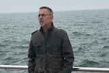A man in a big jacket with his hands in his pockets stands by a sea, looking cold