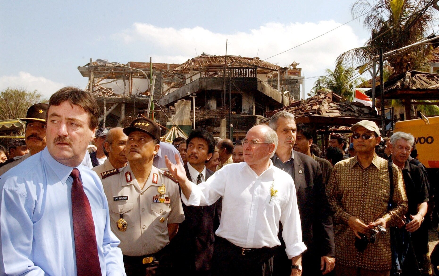 A crowd of men walk through destroyed buildings, Howard at the centre, gesturing to something off camera.