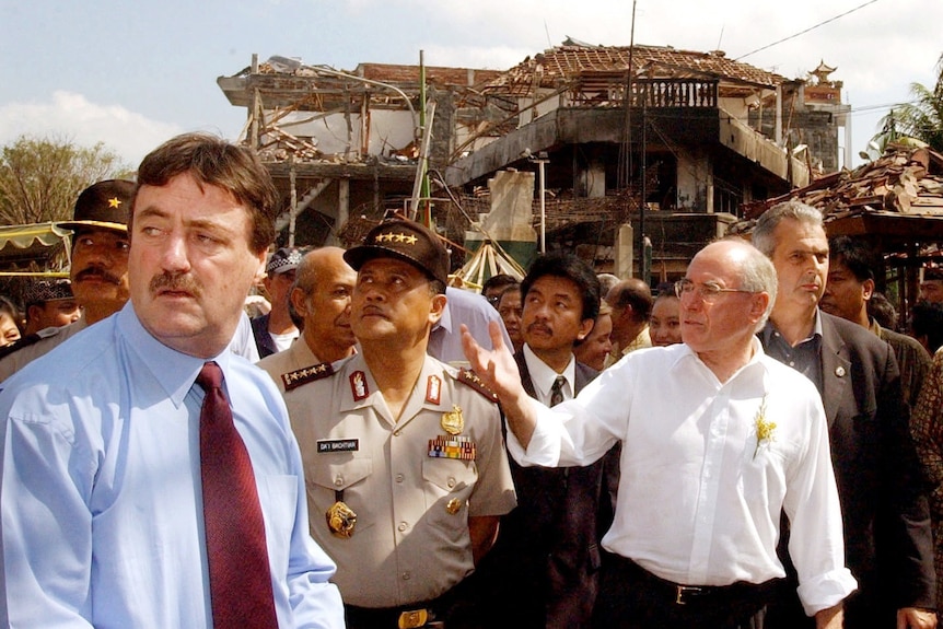 A crowd of men walk through destroyed buildings, Howard at the centre, gesturing to something off camera.