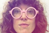 A woman with curly hair is looking straight ahead with large, pink framed glasses on