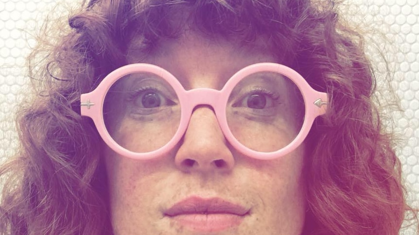 A woman with curly hair is looking straight ahead with large, pink framed glasses on
