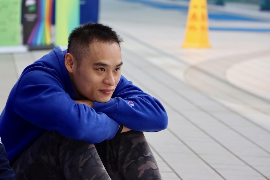 Man sitting on the floor of an indoor swimming pool with his chin resting on his arms that are folded on his knees