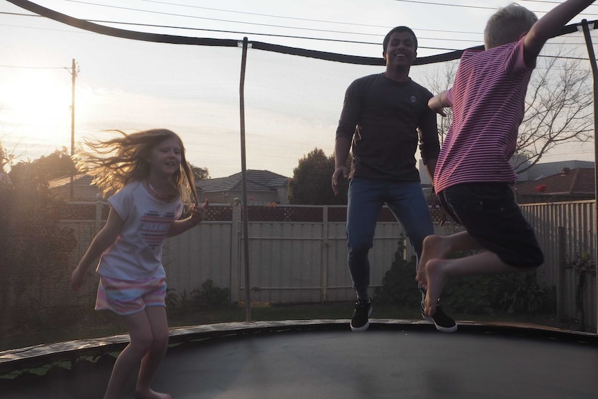 A teenager and two children jumping on a trampoline as the sun sets in the background.