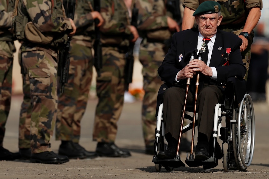 An elderly man sits in a wheel chair and is being pushed past a line of soldiers.