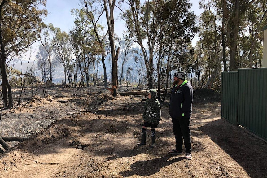 A boy and his dad stand next to each other looking at the smoking burnt trees next to a shed