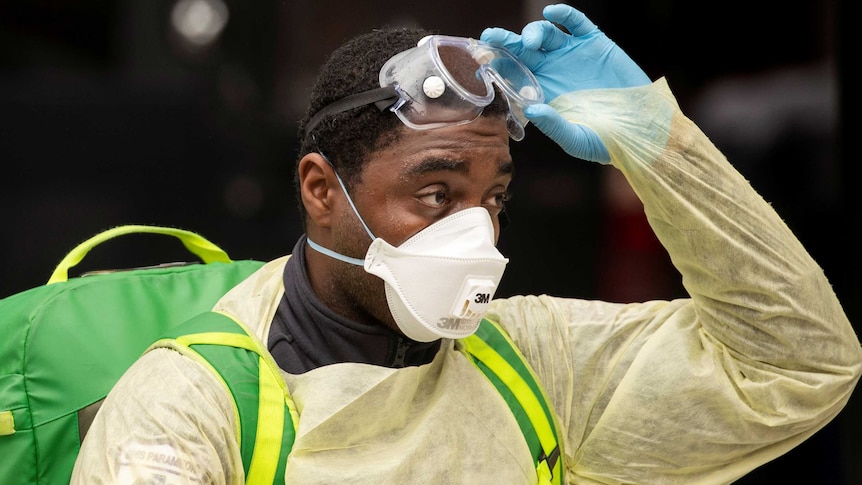 A health care worker in protective gear raises their goggles onto their forehead