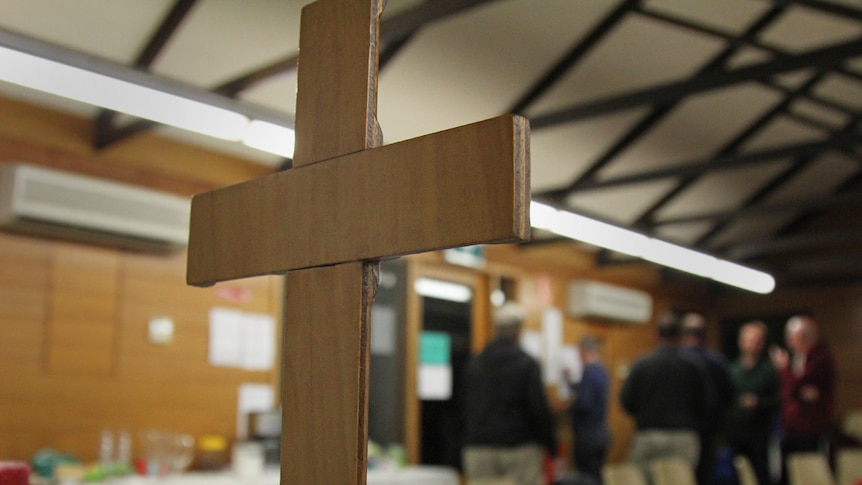 A cross can be seen in focus at the forefront of the photo. People blurred in the background.