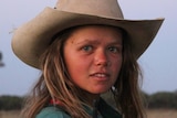 A young woman with a cowboy hat looks directly at the camera