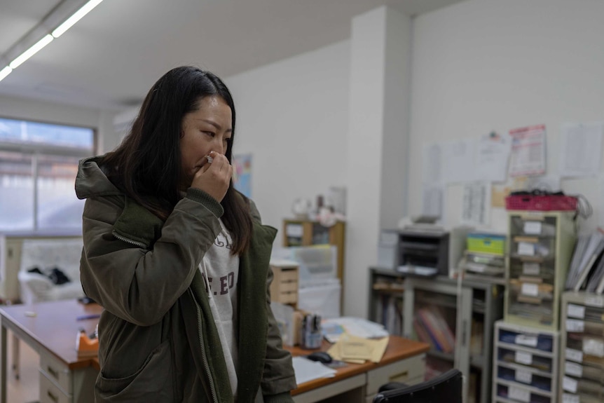 Jianhua Shi wears a khaki coat and grey hoodie with dark hair framing her face. Grief wracks her face as she holds a tissue.