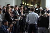 A crowd of journalists and camera operators crowd around a man, standing before several microphones.