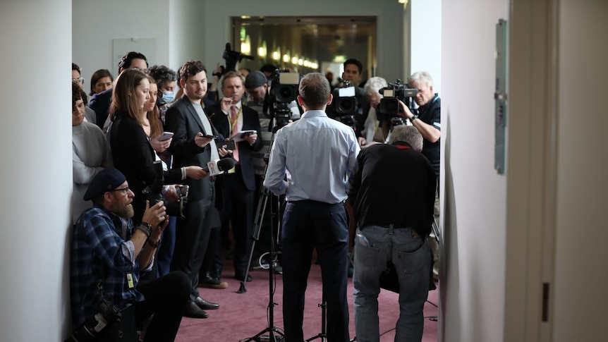 A crowd of journalists and camera operators crowd around a man, standing before several microphones.