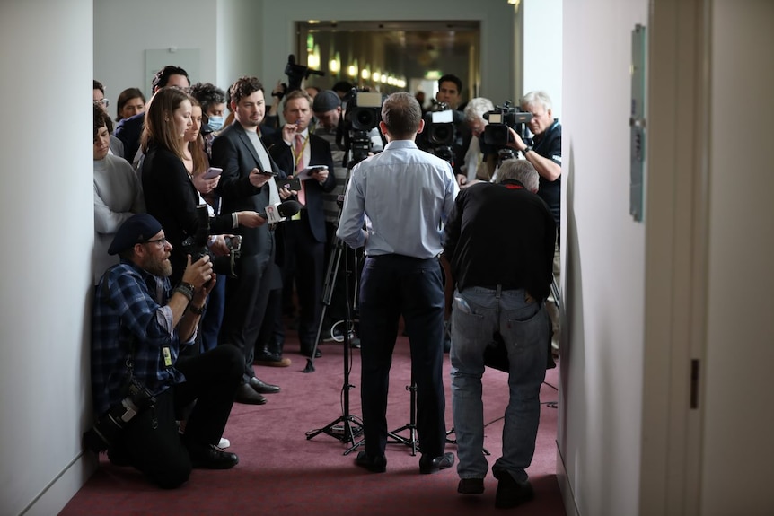 A crowd of journalists and camera operators crowd around a man, standing before several microphones.