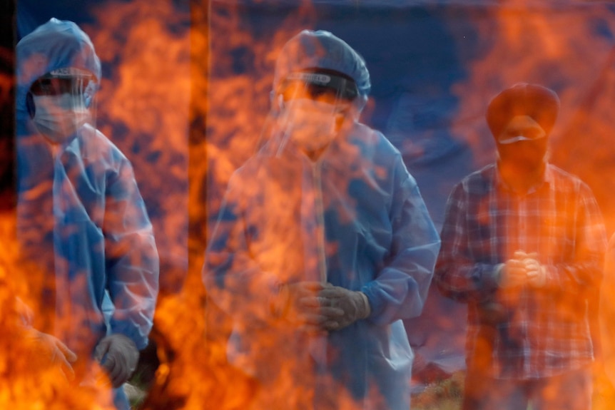 Two men in PPE and one man in street clothes stand obscured behind huge flames