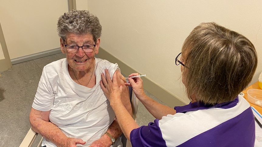 Elderly woman sitting in a chair while woman in a purple shirt puts a surgical needle in her arm.