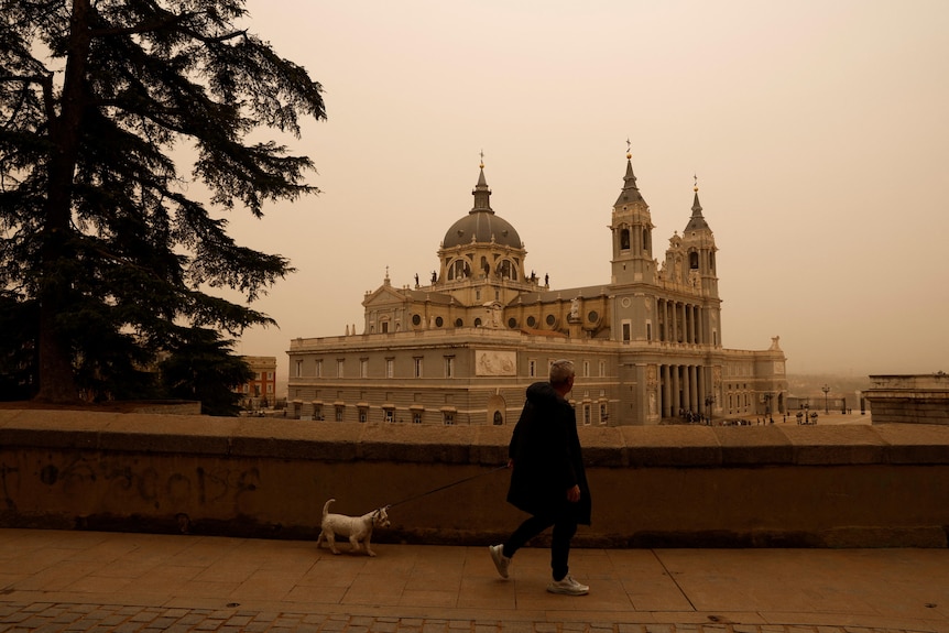 A man walks his dog along a footpath with a cathedral in the background with an orange-tinged sky.