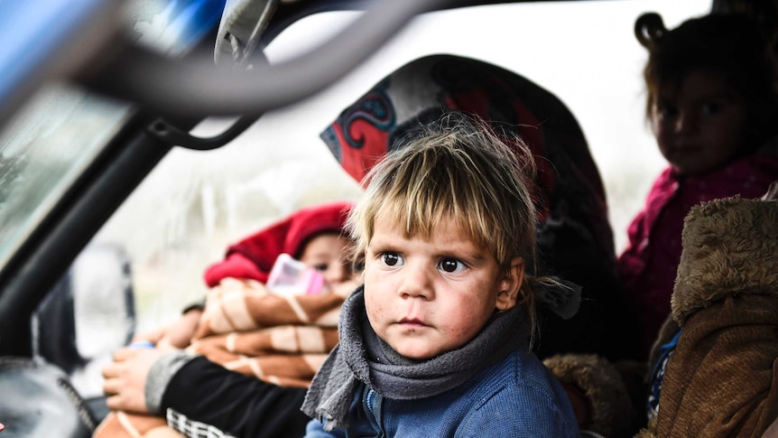 A young, blonde-haired child sits in the front seat of a car fleeing Idlib in Syria.