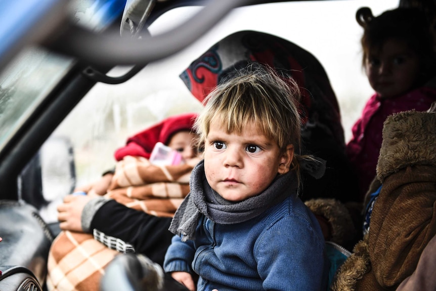 A young, blonde-haired child sits in the front seat of a car fleeing Idlib in Syria.