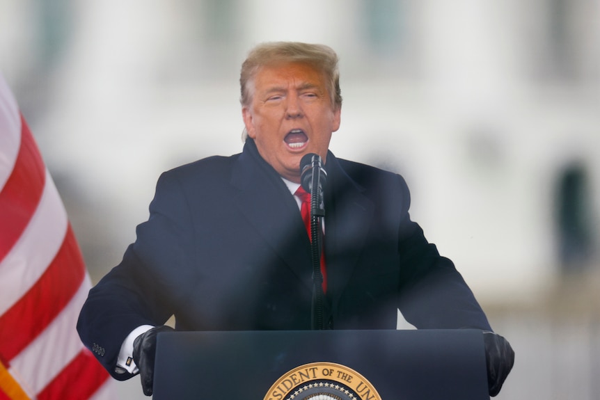 An older man in a black coat and a lot of orange bronzer speaks outside behind a lectern.