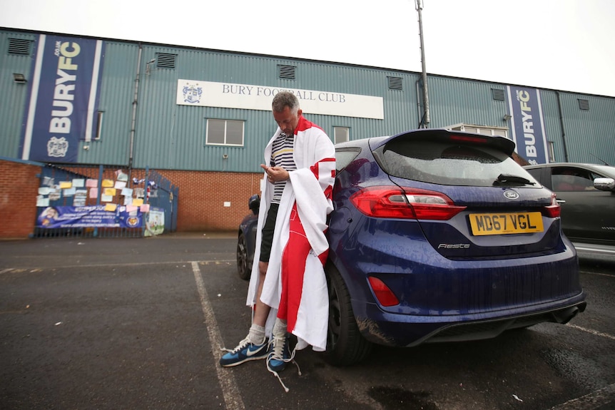 A Bury fan draped in a flag checks his phone leaning against his car outside Bury's stadium