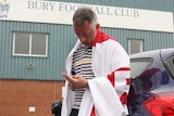 A Bury fan draped in a flag checks his phone leaning against his car outside Bury's stadium