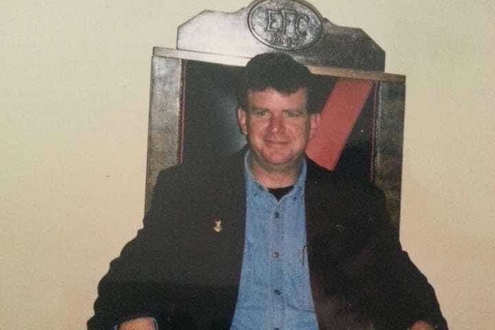 A dark-haired, middle-aged man wearing a dark jacket a denim jeans sits in a grand, throne-like chair.