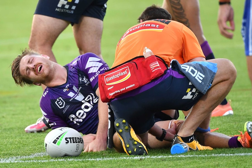 A Melbourne Storm NRL players has an anguished look on his face as he lies on the ground getting treatment on his right knee.
