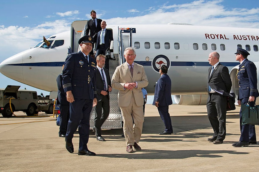 Prince Charles is greeted by Australian airforce personnel as he walks off a white plane on a sunny day.