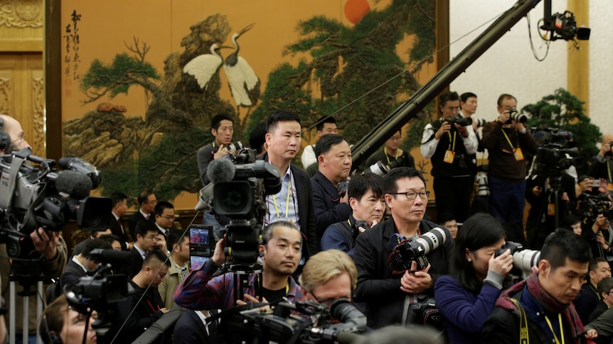 A group of journalists wait in a tight group with cameras ready for a meeting in a formal hall.