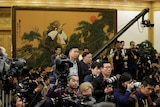 A group of journalists wait in a tight group with cameras ready for a meeting in a formal hall.