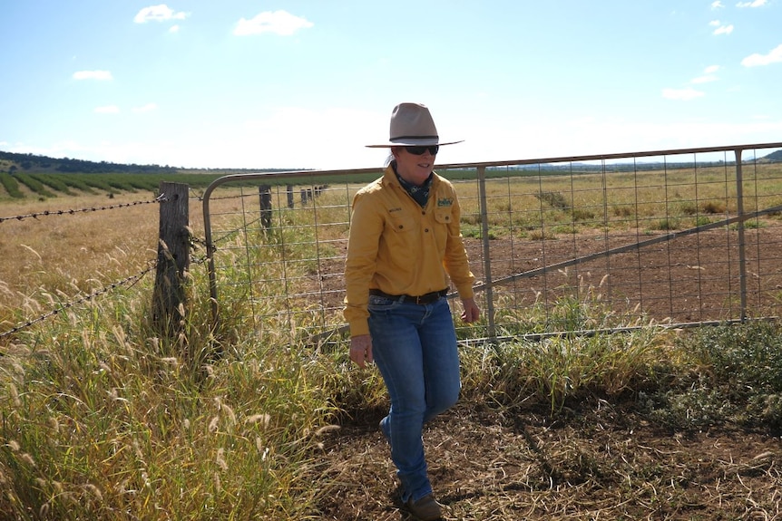 A woman in a bright button up work shirt in jeans walks away from a wire gate in a dry looking paddock  
