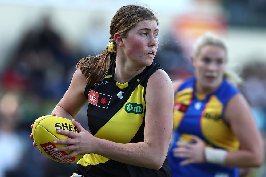 A Richmond AFLW player holds the ball in two hands against West Coast.