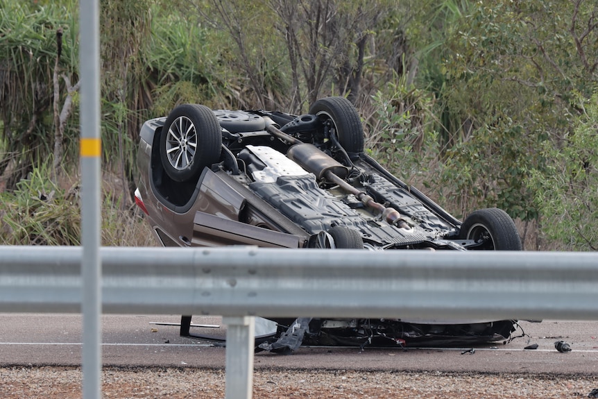 A crashed car rested on its roof on the side of a highway, in front of scrub. 