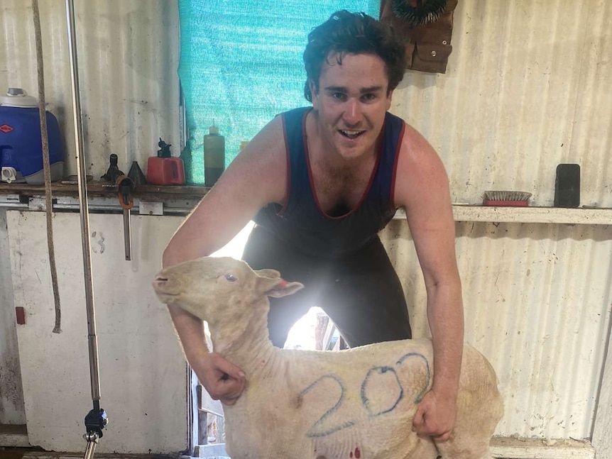 A young man in a singlet smiles while he holds a sheep with the number 200 painted across its body.