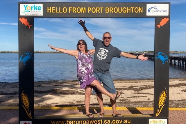 Kellie and Aaron pose in a tourism frame in front of a beach that reads 'hello from Port Broughton'.