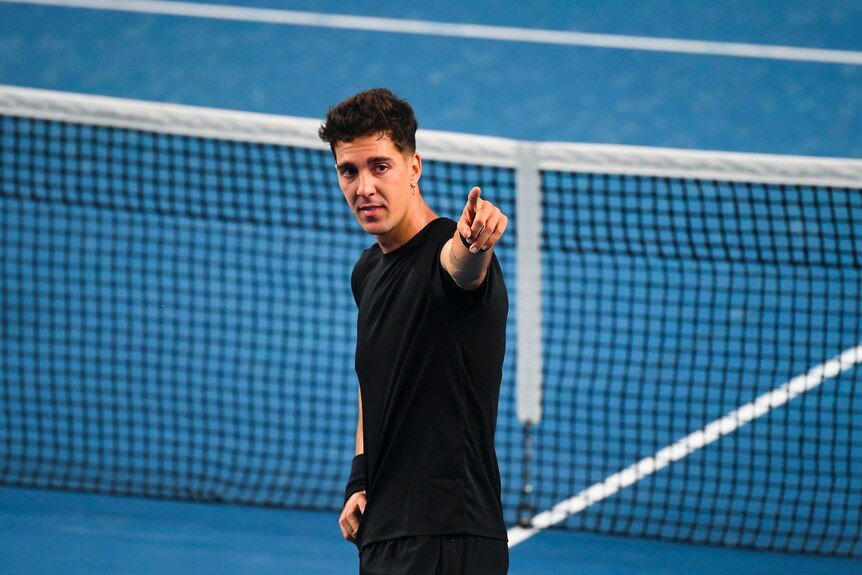 Dark haired man in dark shirt points with tennis net in background