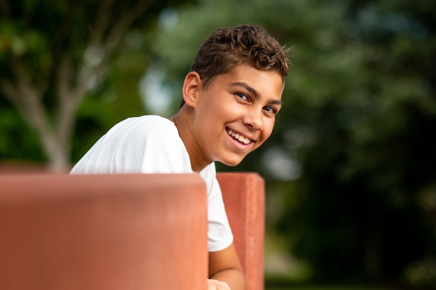 a young boy in a white shirt leans on a park bench and smiles at the camera.
