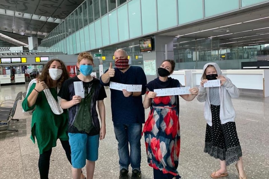 Two women, a man and two kids wearing masks hold tickets up to the camera as they wait at an airport.