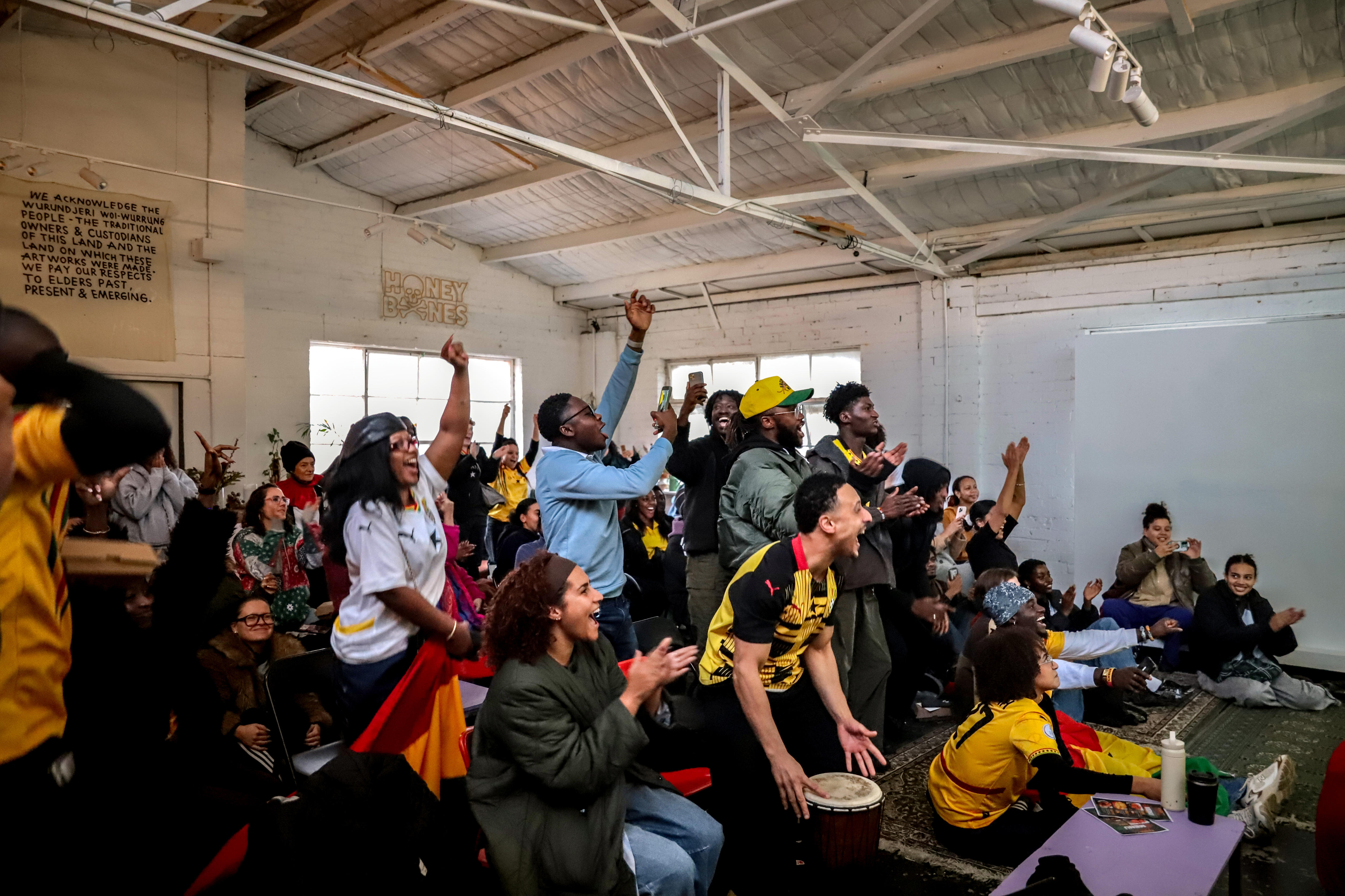 A room full of people, some wearing red, green and yellow, clap and raise their arms as they look forward.