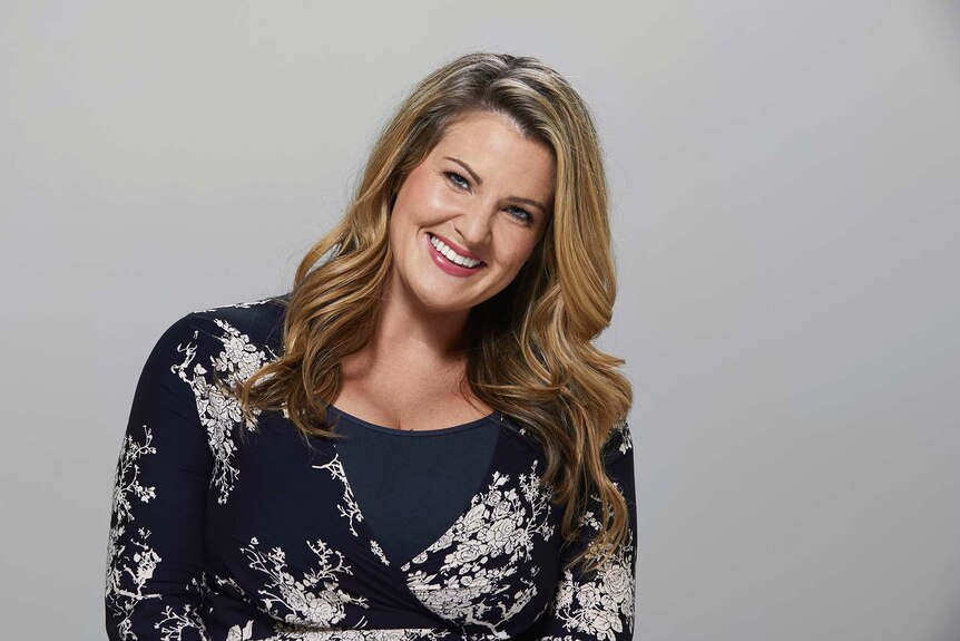 Comedian Nikki Britton photographed in a studio, smiling at the camera and wearing a floral wrap dress. She goes on dates alone.