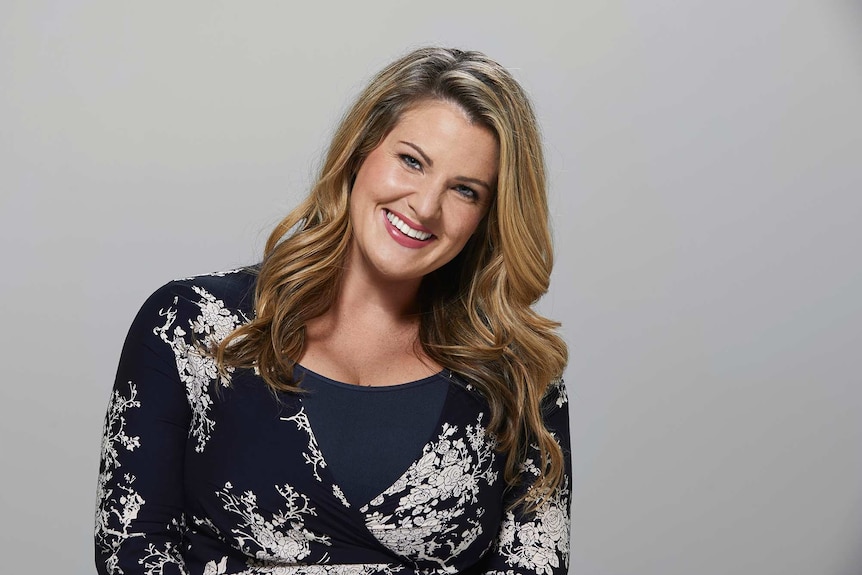 Comedian Nikki Britton photographed in a studio, smiling at the camera and wearing a floral wrap dress. She goes on dates alone.