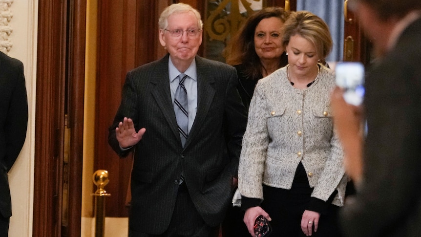 A man waves his hand while wearing a suit. A woman stands next to him.
