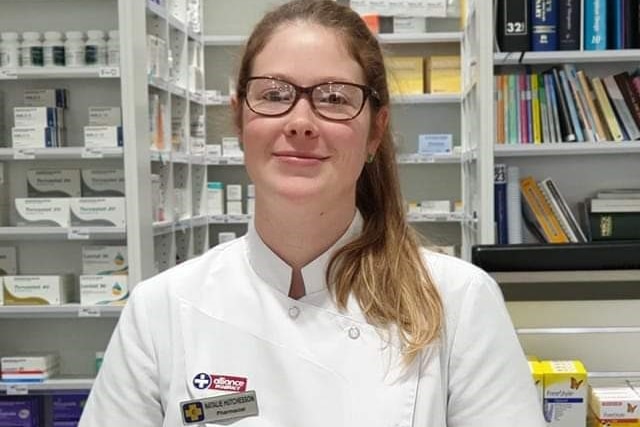 A woman with glasses in a white uniform smiles at the camera with stacks of medication in the background