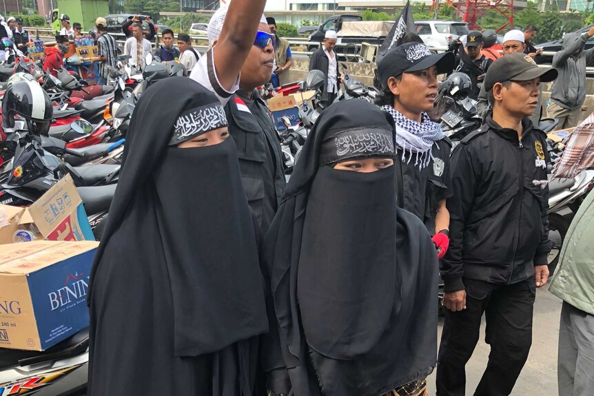 Two women wearing niqab and covering their faces stand in front of a group of protesters in Indonesia