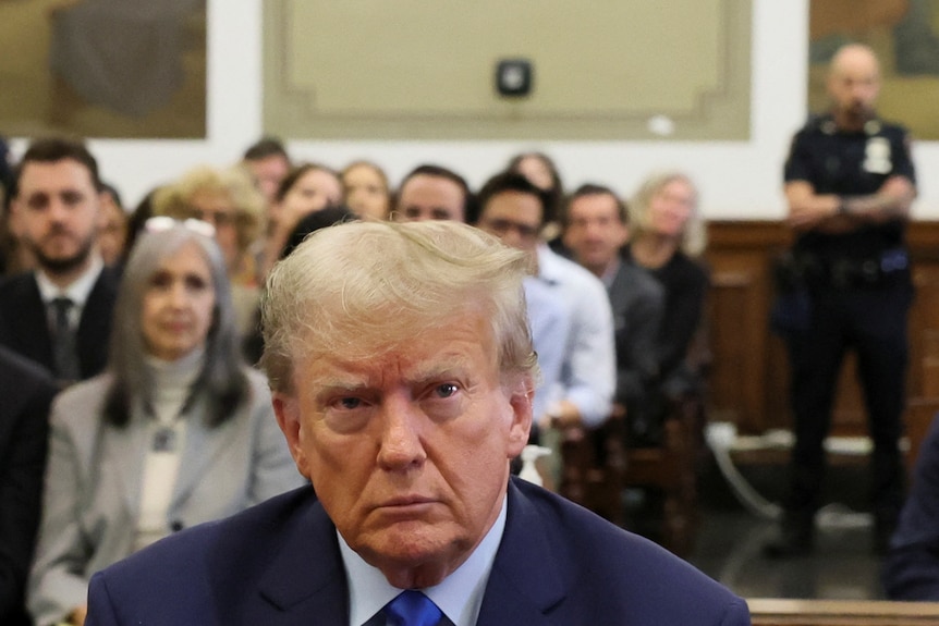 Donald Trump sits in court and looks straight ahead. The gallery behind him is full of people.